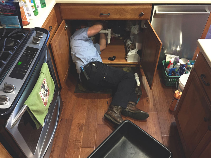 plumber lying inside kitchen cabinet performing under sink pipe repair