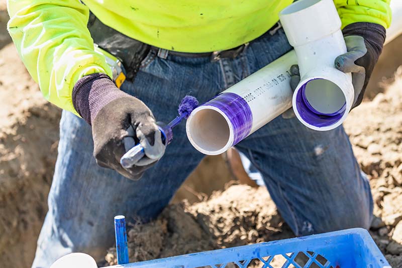 Plumber Applying PVC Primer to Sewer Pipe in Trench plumber applying purple pvc primer to sewer pipe fitting in excavated trench