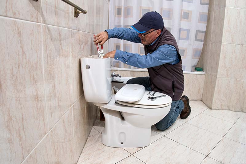 Plumber Repairing Toilet Tank in Tiled Bathroom plumber kneeling to repair toilet tank in tiled bathroom with tools on seat